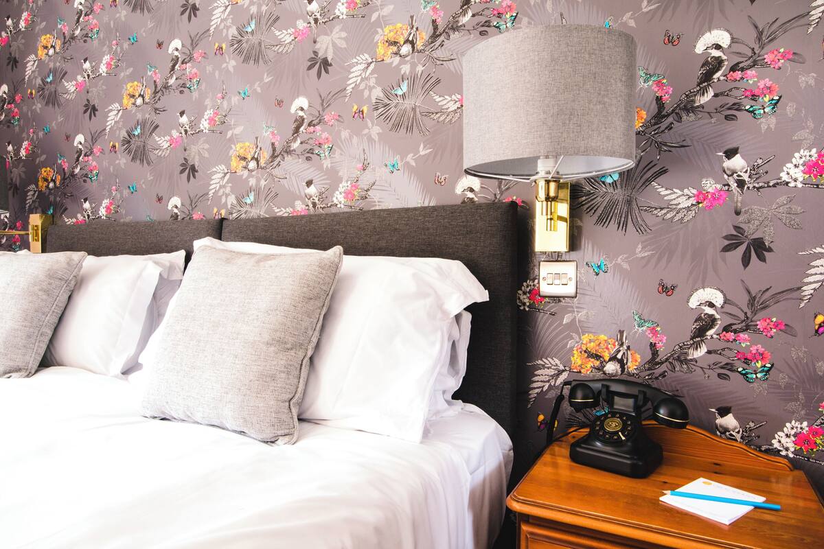 A neatly made bed with white linens and grey pillows, a wooden nightstand with a telephone and a notepad, and a wall with floral wallpaper.