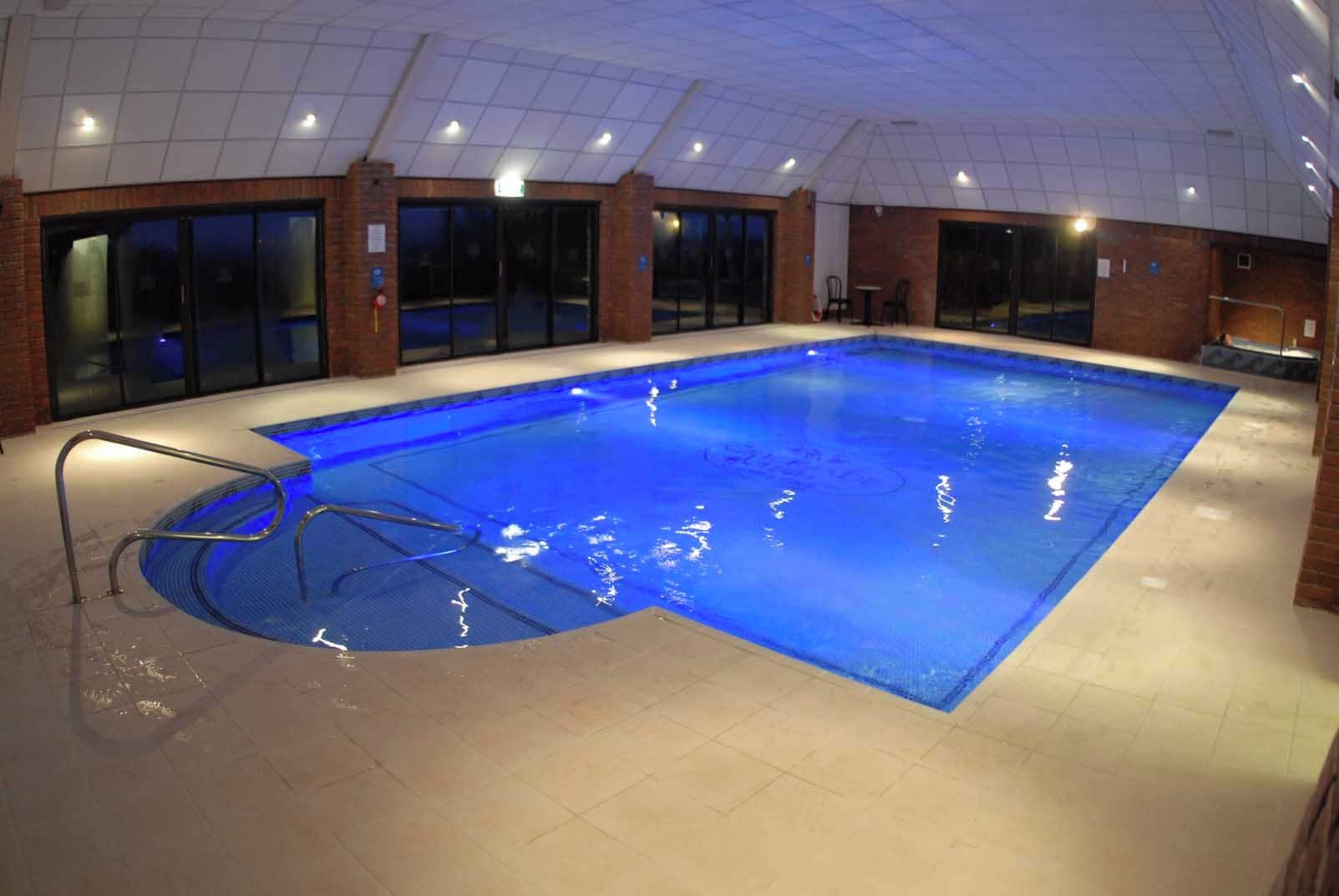 Una piscina interior con superficie de agua cristalina, rodeada de suelo y paredes con mosaicos, y equipada con una escalera de metal.