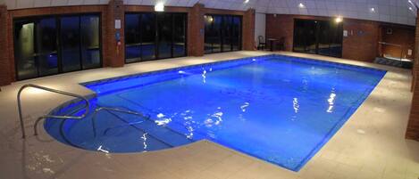 Una piscina interior con superficie de agua cristalina, rodeada de suelo y paredes con mosaicos, y equipada con una escalera de metal.