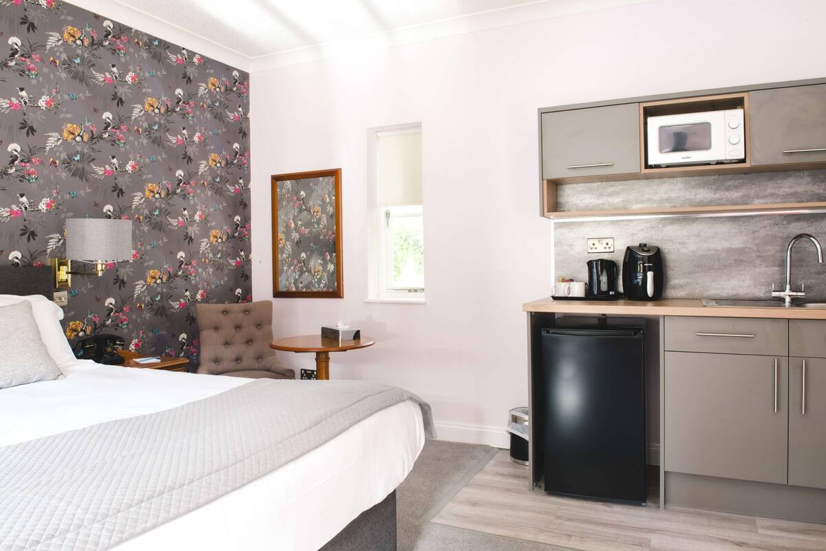 A compact hotel room with a bed, a small desk, a chair, a microwave, a coffee maker, and a refrigerator.