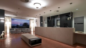 A modern living room with a sofa, a coffee table, and a dog. There is a large window with curtains, a flat-screen TV, and a kitchen area with a counter and cabinets.