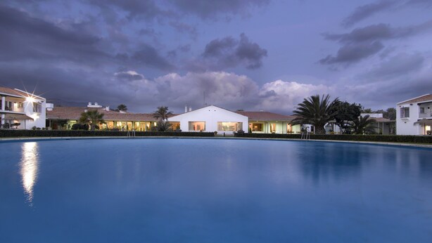 A pool with a view of a white building with a tiled roof and multiple balconies.
