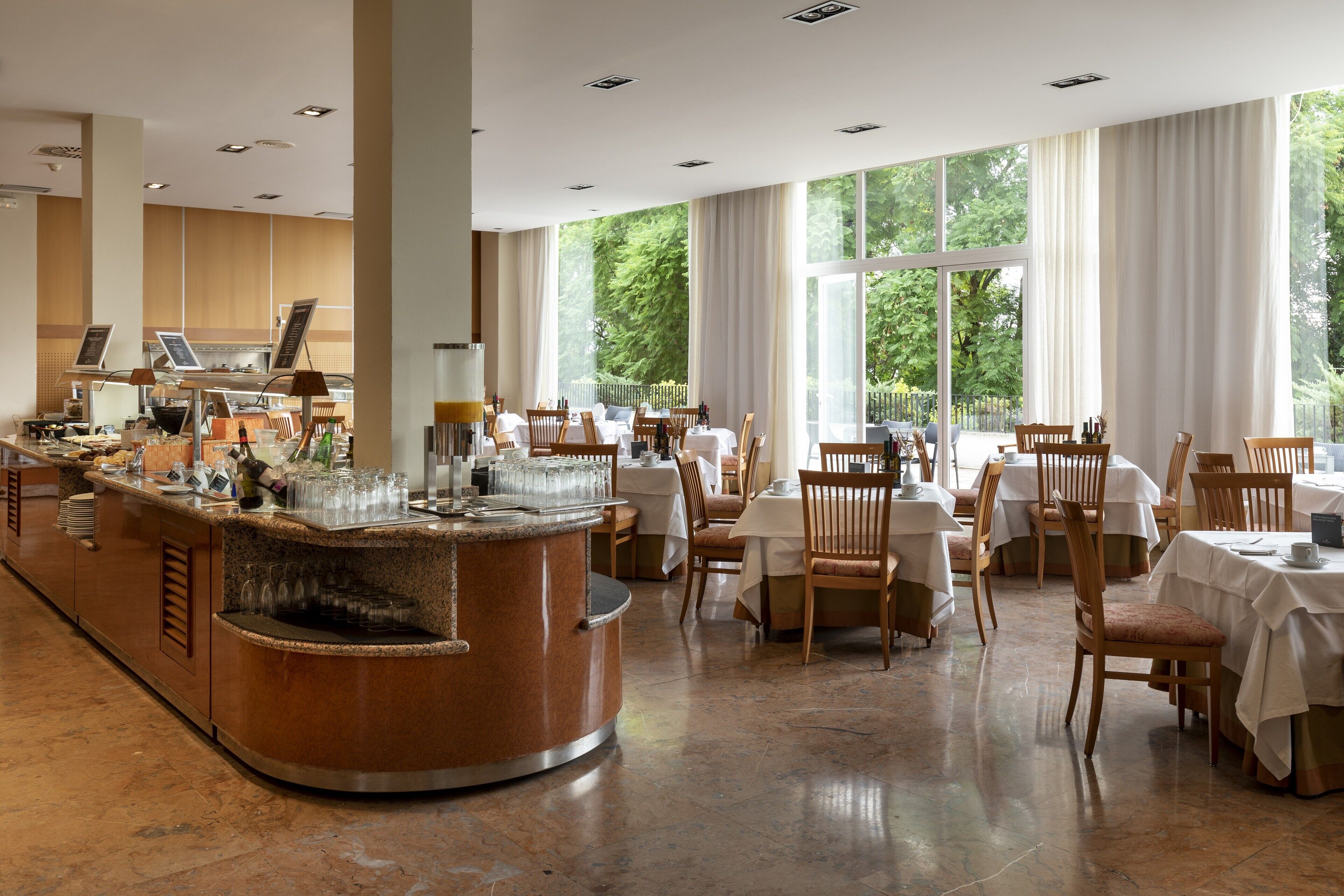 A spacious restaurant with a central bar, multiple dining tables, and large windows offering a view of the outdoors.