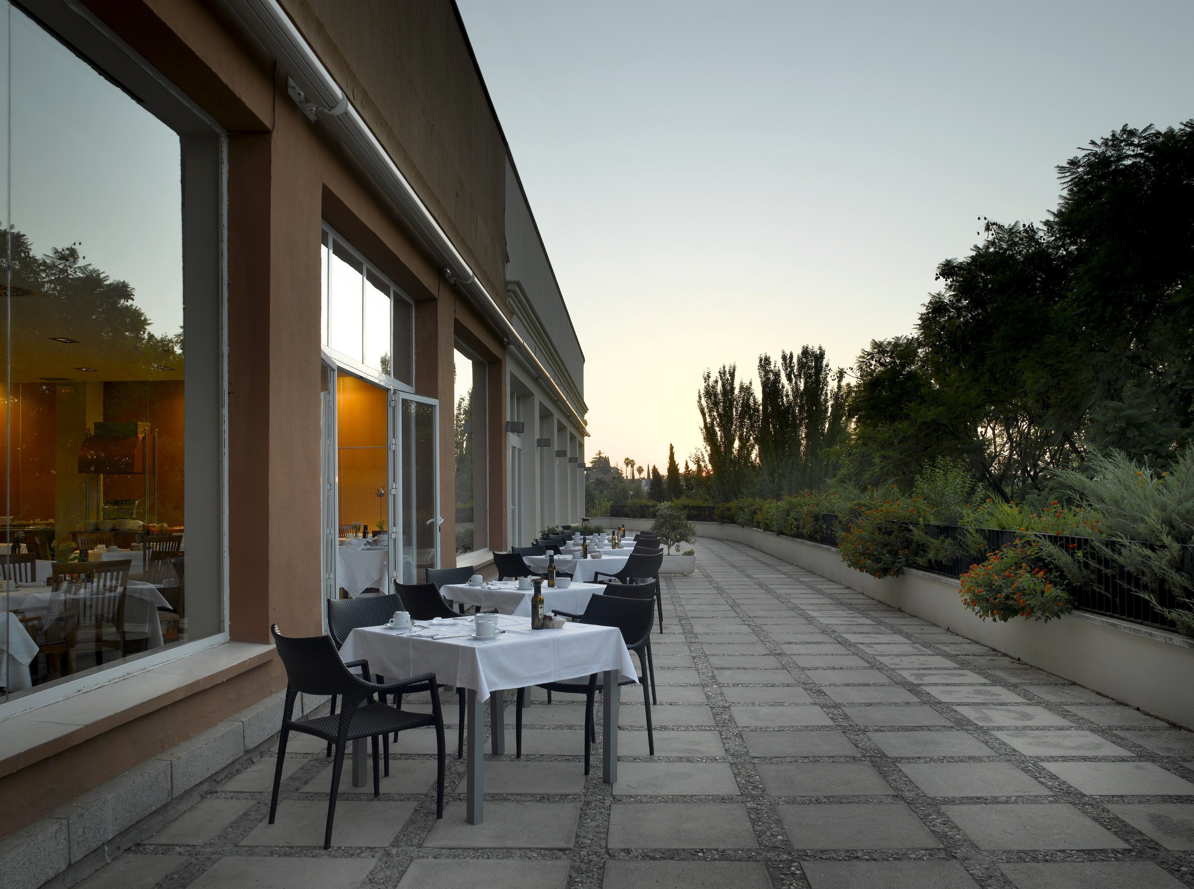 Un área de comedor al aire libre con mesas y sillas, un edificio con grandes ventanales y un sendero bordeado de follaje.