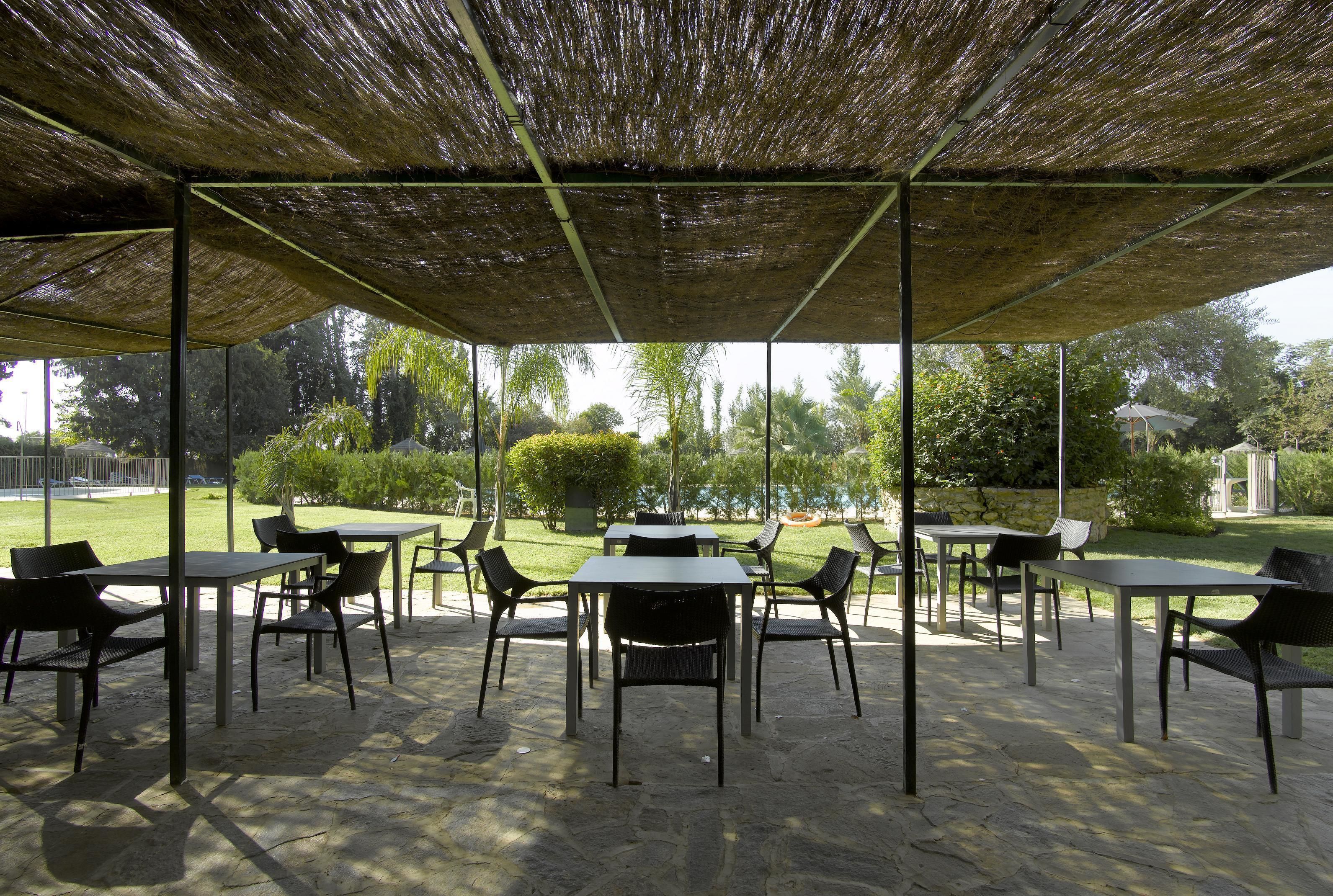 Zona de terrassa a l'exterior amb taules i cadires, coberta per un teulat de palla i envoltada de vegetació.