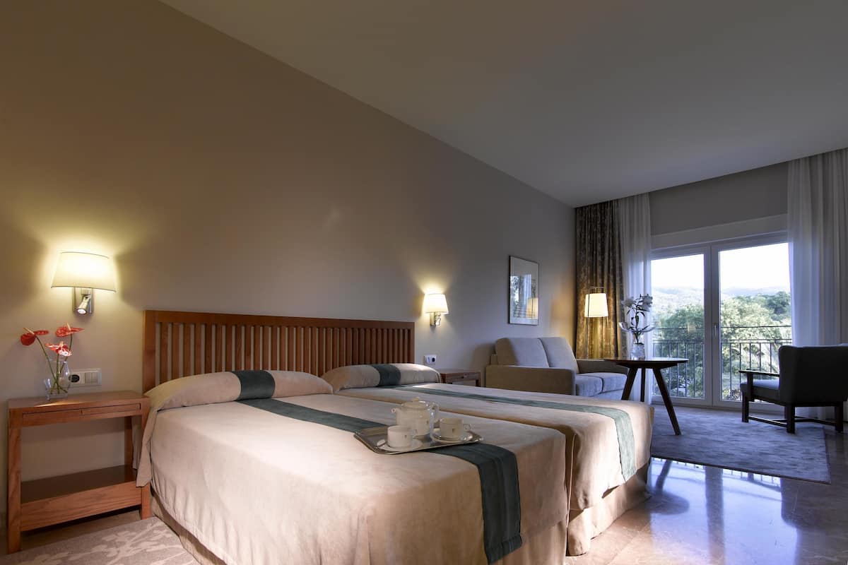 A hotel room with two beds, a sitting area with a chair and a table, and a balcony with a view.