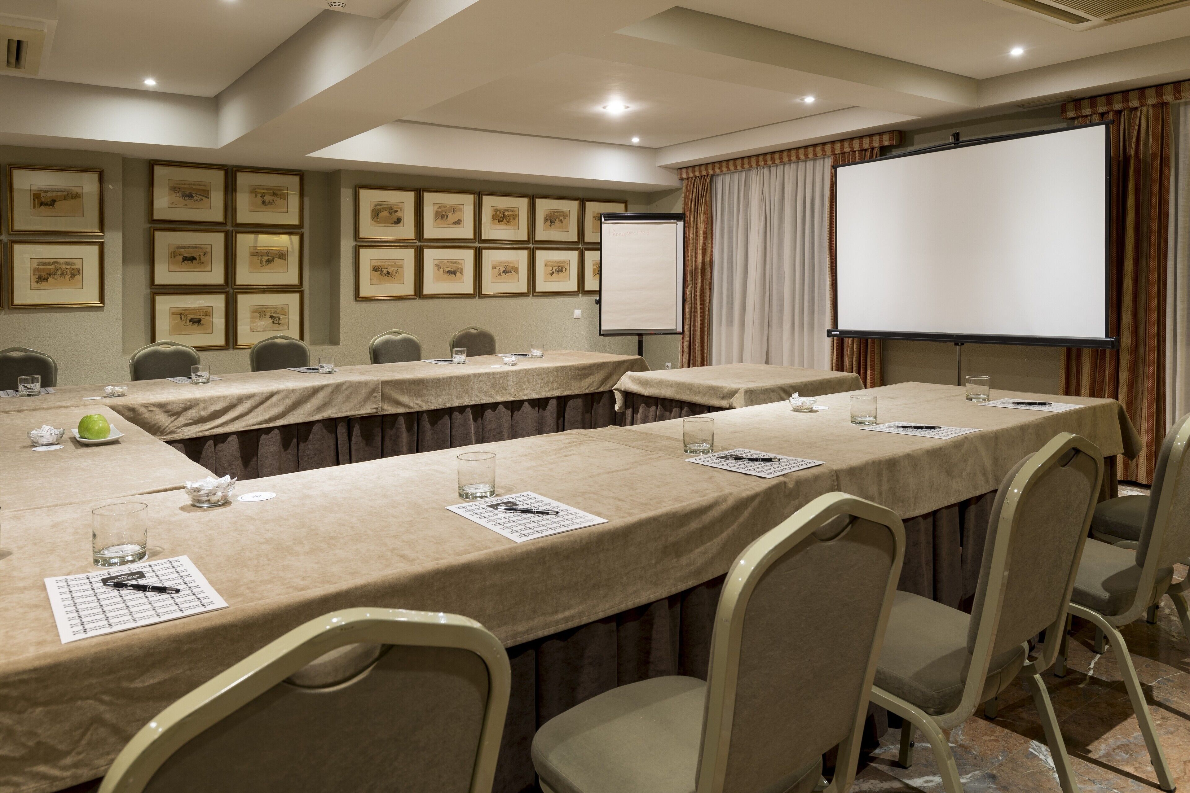 Un salón de conferencias con mesas largas, sillas, una pantalla de proyector y cuadros en la pared.