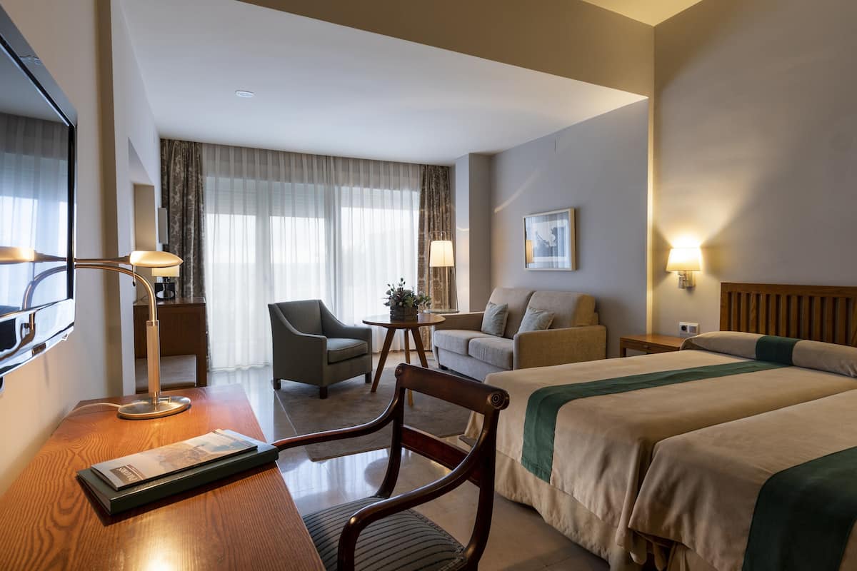 A hotel room with two beds, a sofa, a chair, a desk with a lamp, a TV, and a window with curtains.