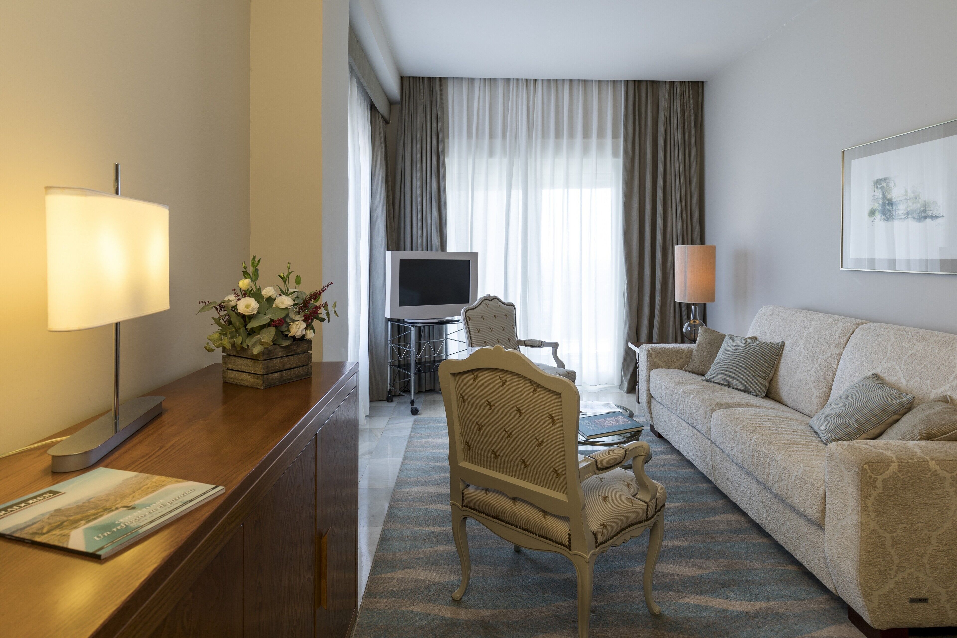Habitación de hotel con sofá, una silla, televisor, lámpara y un arreglo floral.