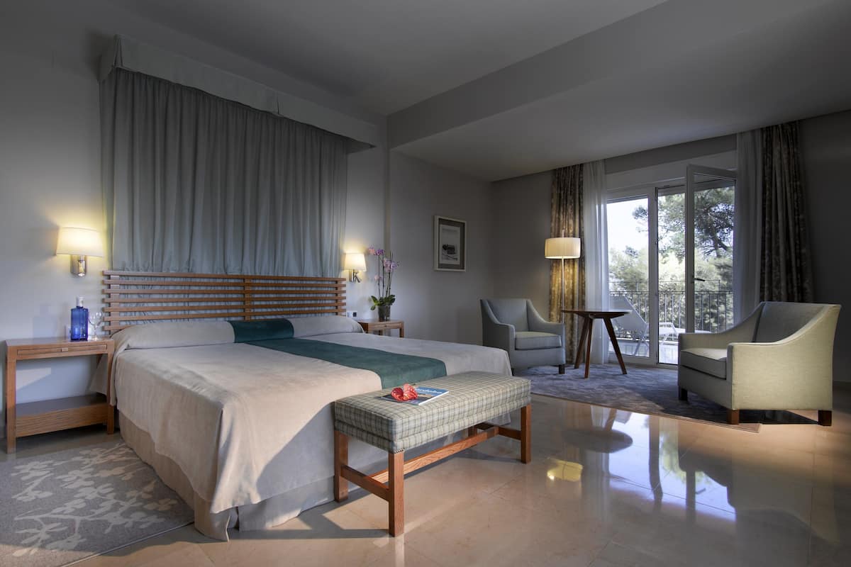 A modern hotel room with a large bed, a bedside table, a chair, a small table, and a view of the outdoors.