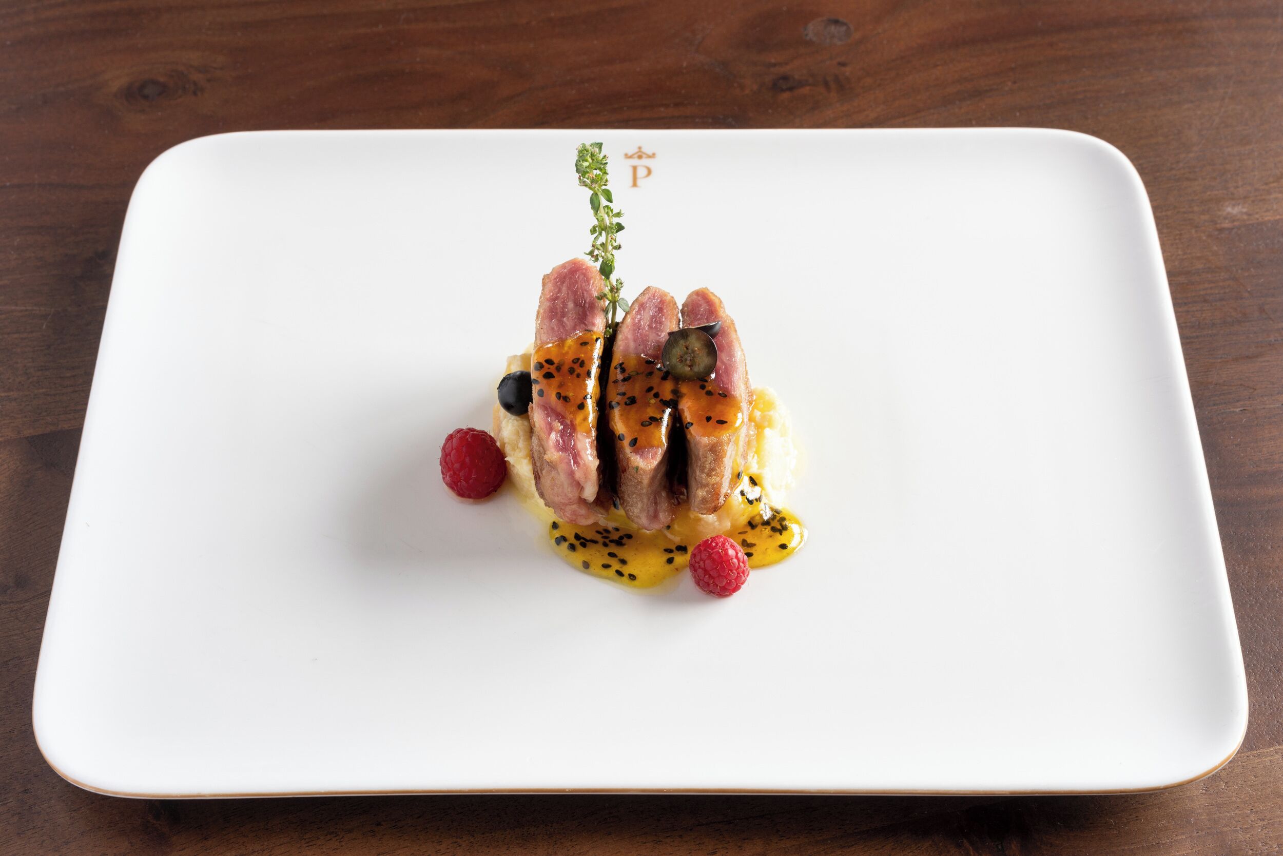 A white square plate with a dish of food, including meat and berries, on a wooden table.