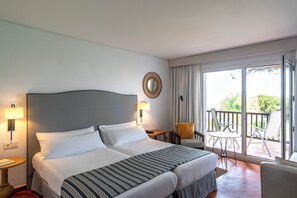 A hotel room with two beds, a balcony with chairs, and a view of greenery. - Parador De Mazagon (Moguer)