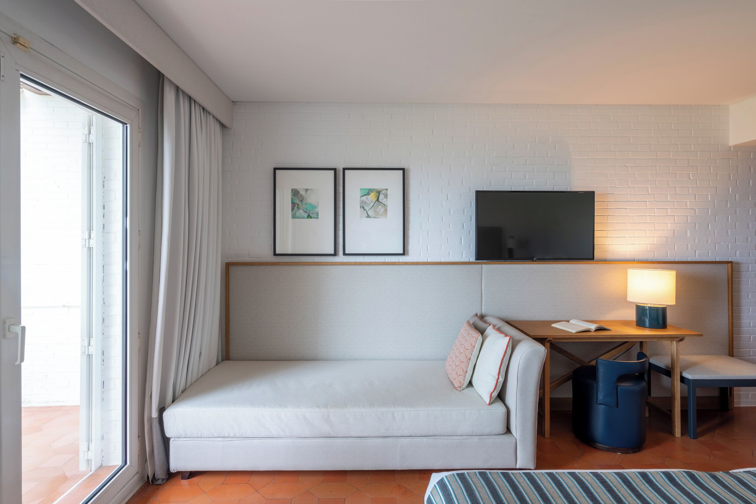 A modern hotel room with a white brick wall, a sofa, a desk with a lamp, and two framed pictures.