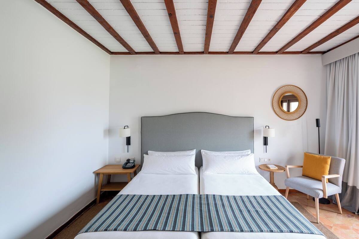 Habitación de hotel con una cama grande, dos mesitas de noche, una silla, un espejo redondo y un techo con vigas de madera.