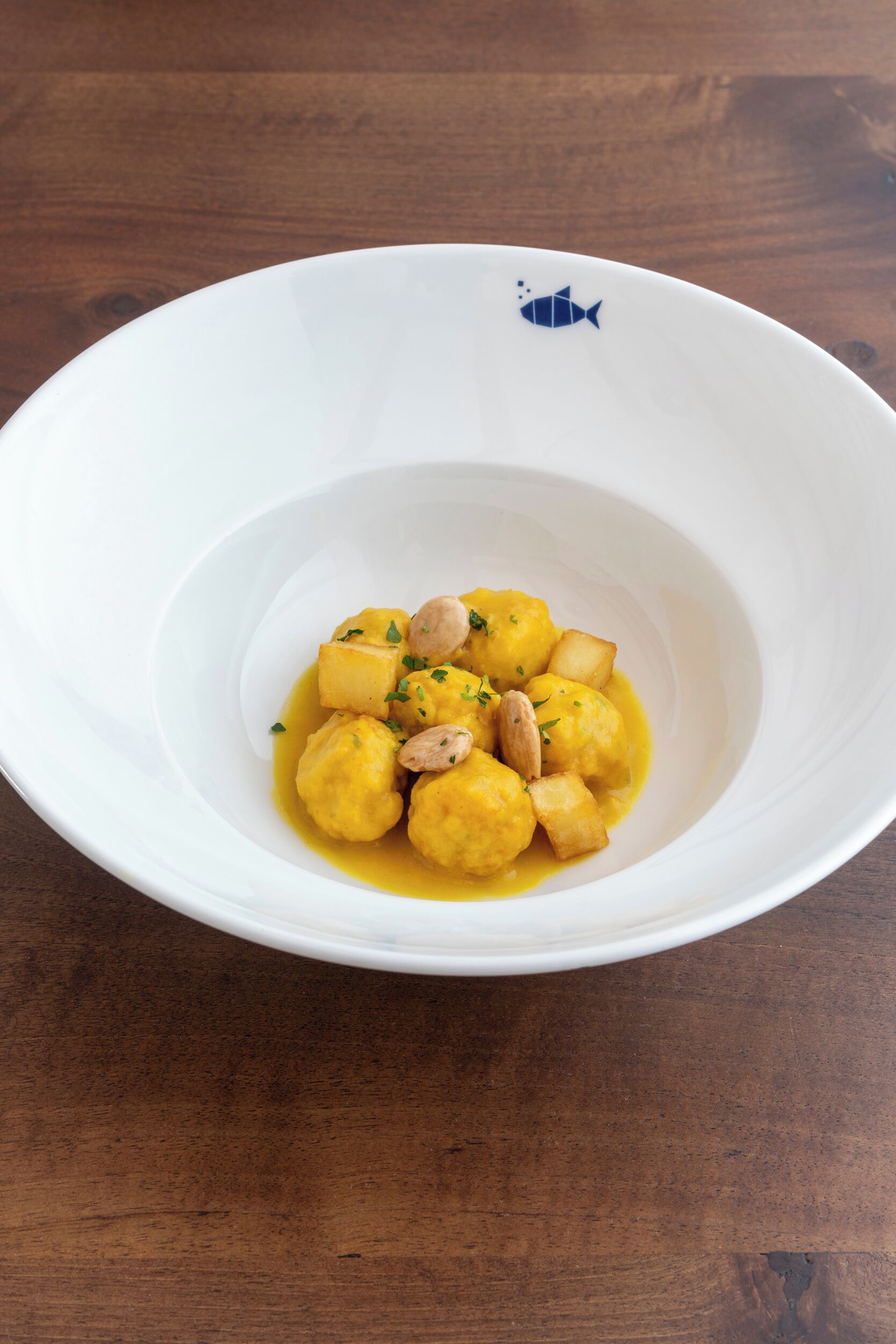 A white bowl with a fish design on the rim, containing a dish of yellow-colored food garnished with herbs and nuts.