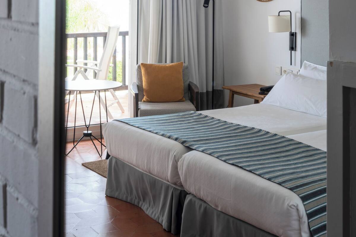 Habitación de hotel con cama, una silla, una mesa, una lámpara y un balcón con vistas.