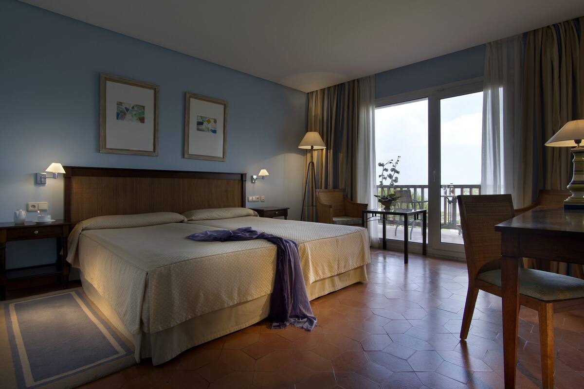 Habitación de hotel con una cama grande, dos mesitas de noche con lámparas, una silla, una mesita pequeña con una lámpara y un balcón con vistas.