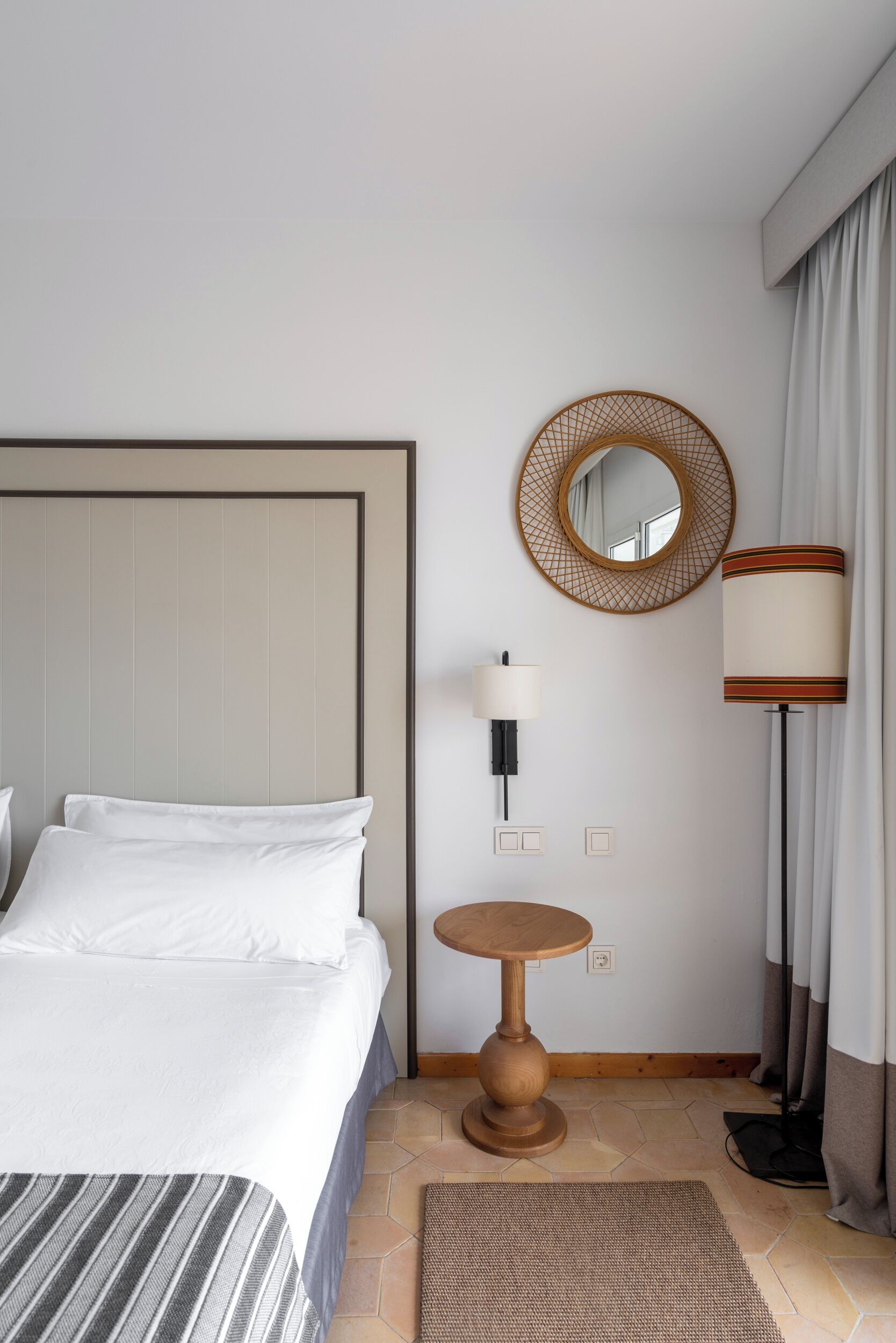A hotel room with a large bed, a round mirror, a bedside table, a lamp, and a view of the outdoors.