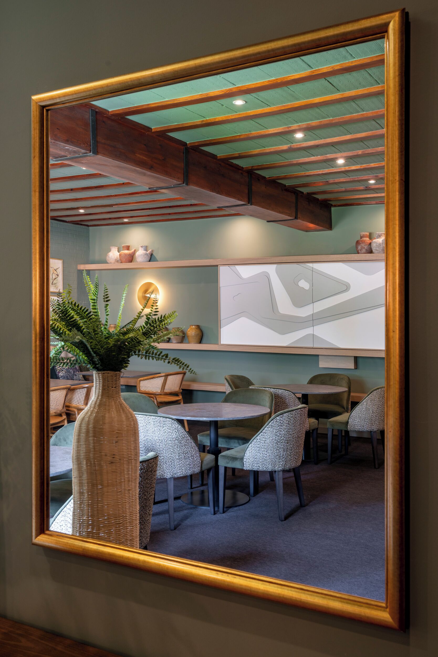 A dining area with a large mirror reflecting the space, including a round table and chairs, a vase with greenery, and shelves with decorative items.