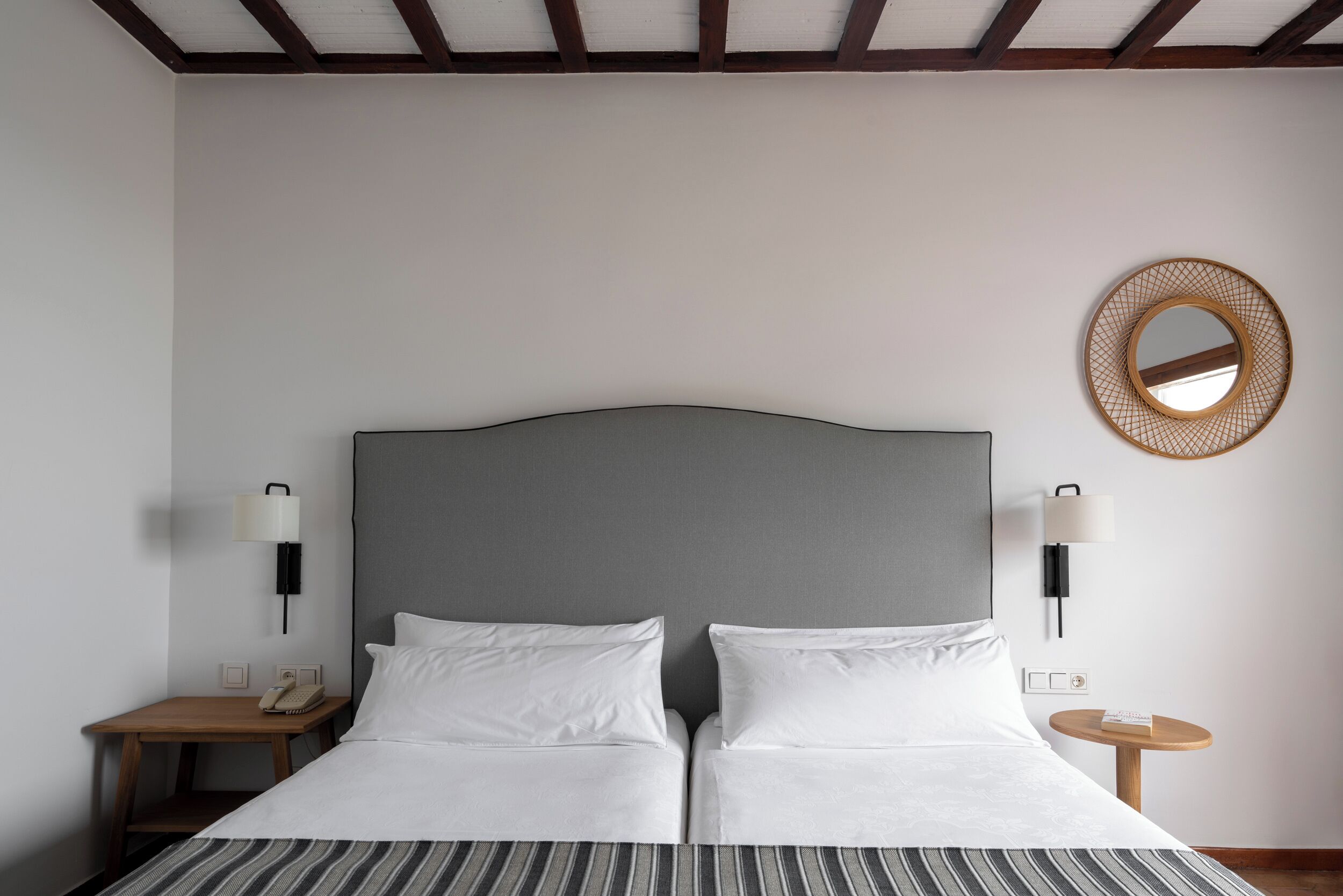 A double bed with a grey headboard, two bedside tables with lamps, a round mirror on the wall, and a striped area rug.
