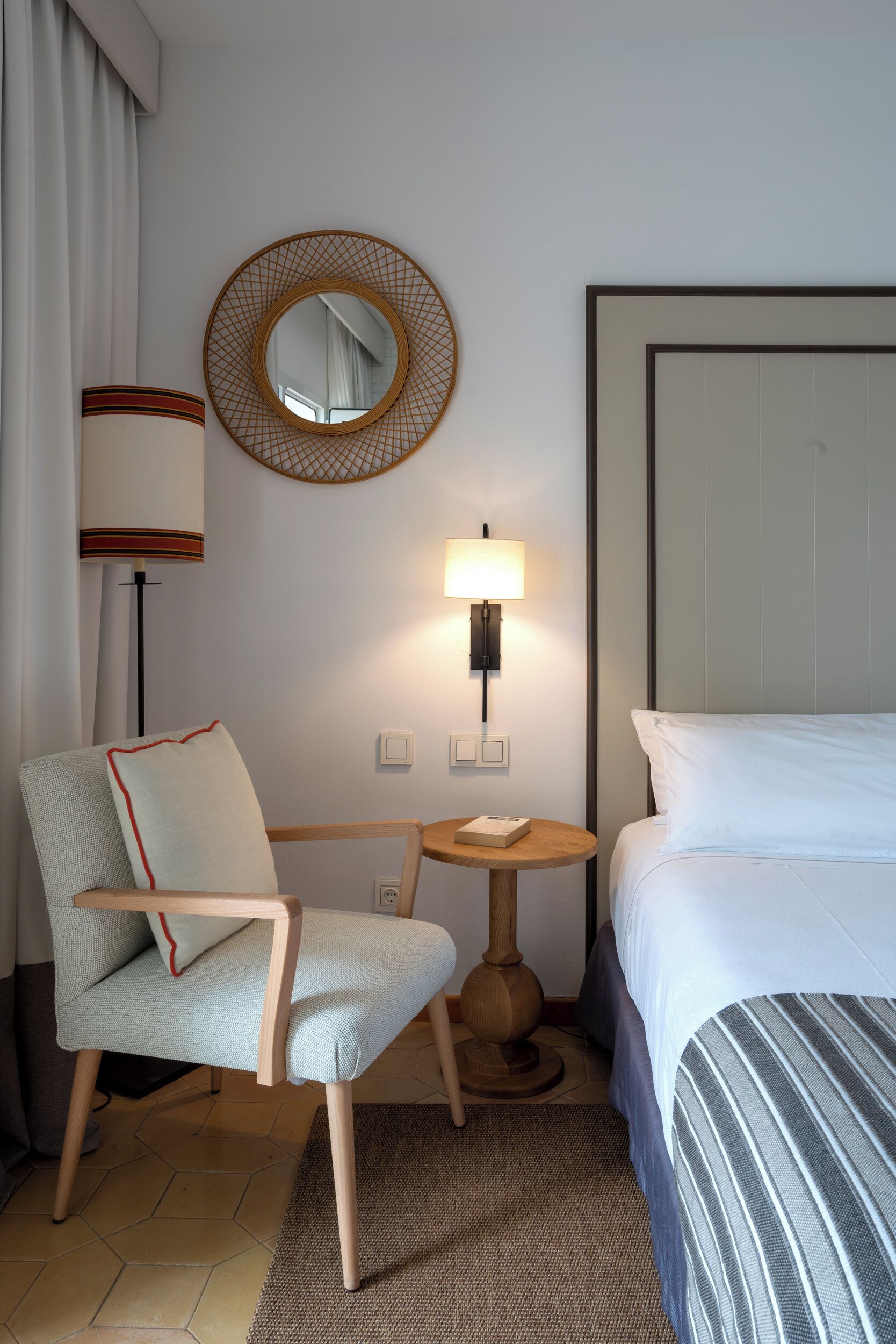Hotellihuone, jossa on sänky, tuoli, pyöreä peili, lamppu ja pieni pöytä.
