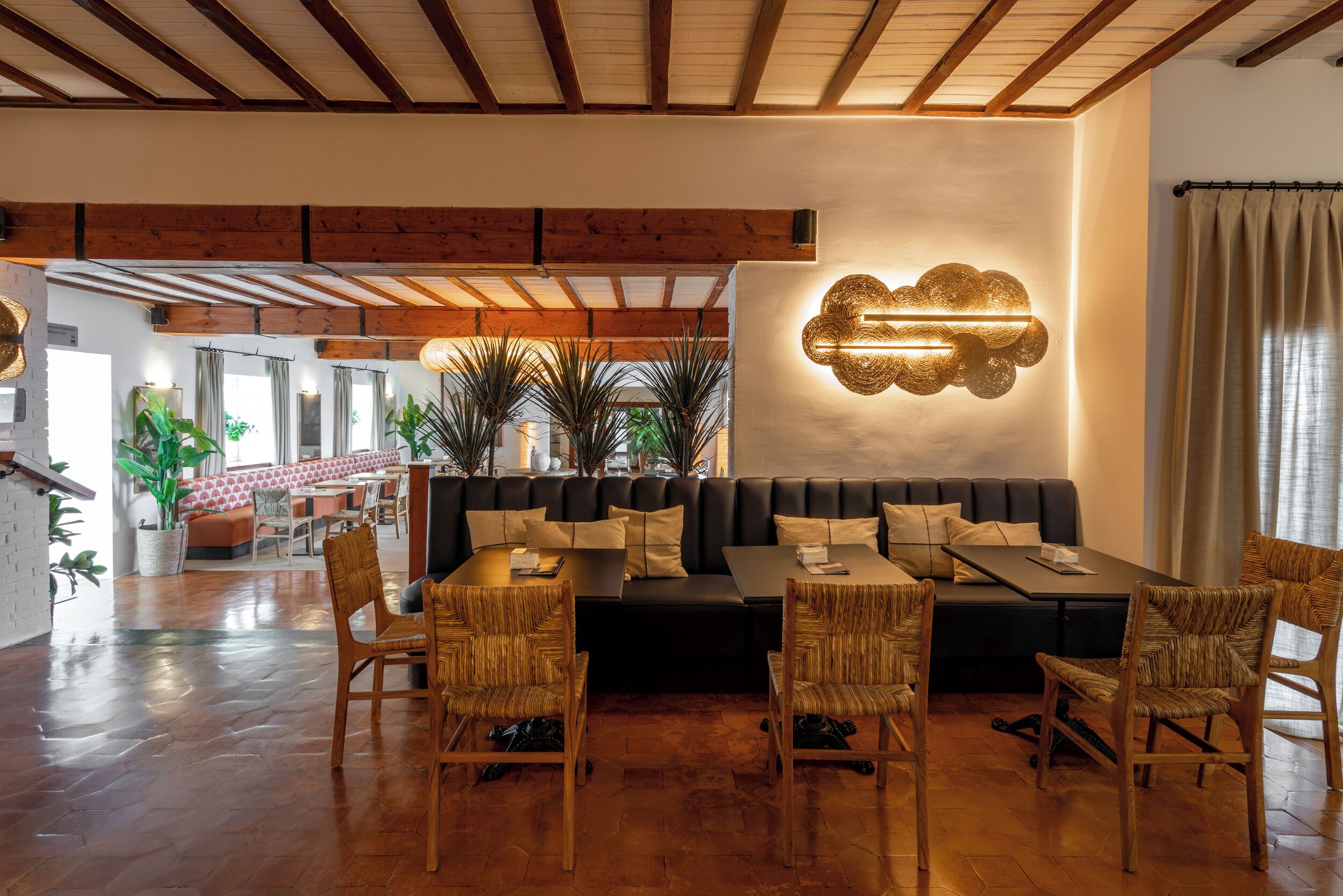 A spacious lobby with a black sofa, wooden chairs, and a table. There is a wall-mounted light fixture and a plant in the background.