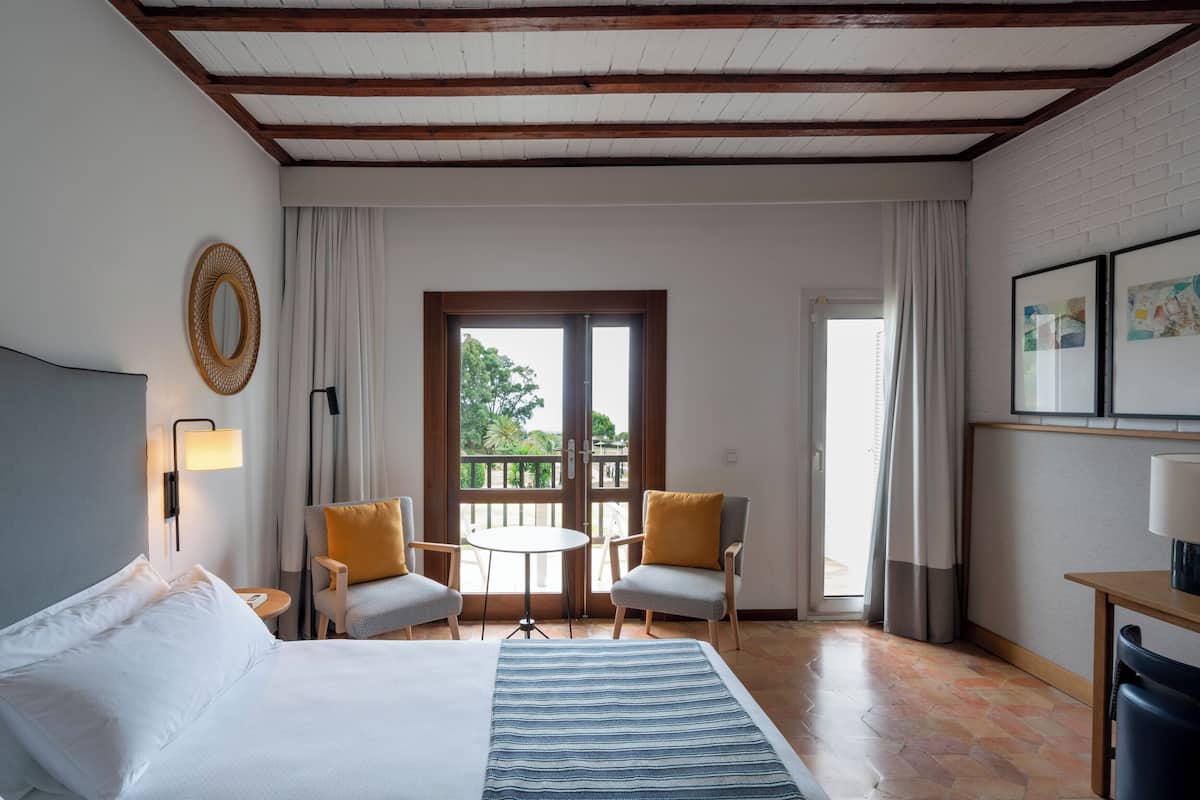 Habitación de hotel con cama, dos sillones, una mesita, lámpara, espejo y ventana con cortinas.