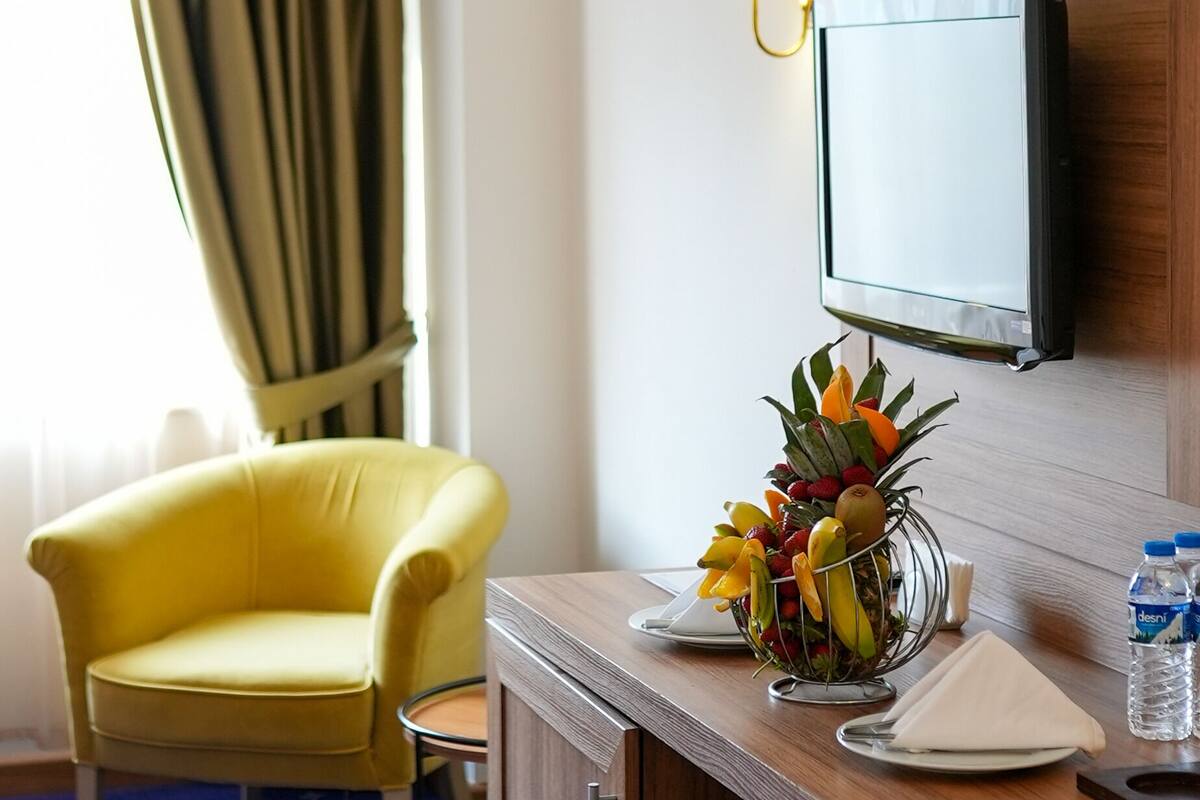 Ein Hotelzimmer mit Holztisch, gelbem Sessel, Wandfernseher, Lampe, Obstschale und Wasserflasche.