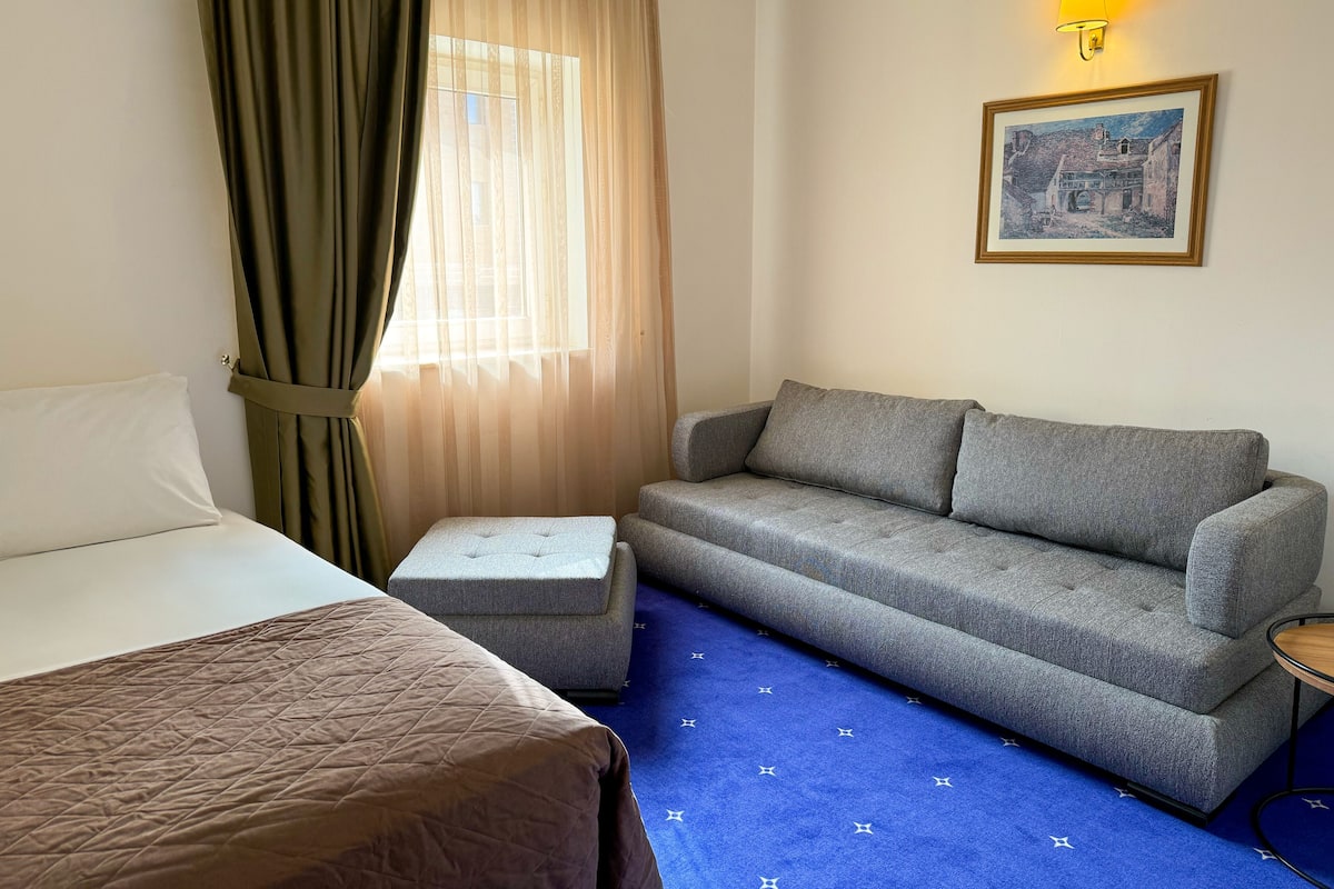 Ein Hotelzimmer mit einem Bett, einem grauen Sofa, einem kleinen Hocker, einem blauen Teppich mit Sternenmuster, einem Fenster mit transparenten Vorhängen und einer Wandleuchte.