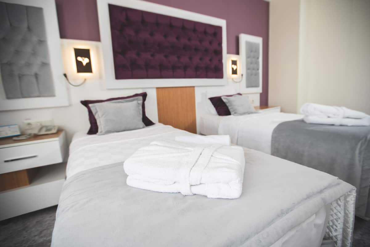 Habitación de hotel con dos camas, cada una con una almohada y una toalla doblada. Hay una mesita de noche con una lámpara y un teléfono.