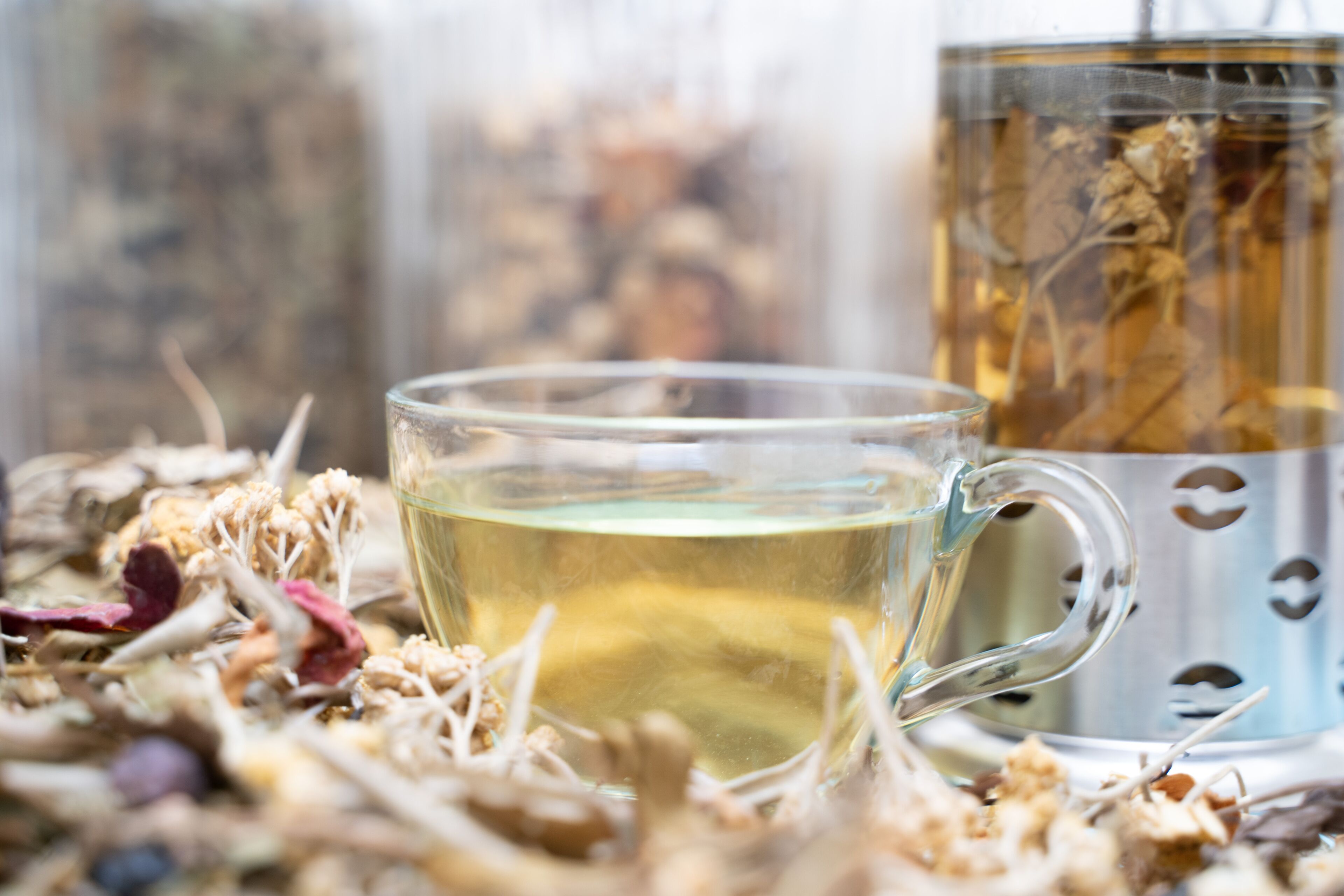A clear glass cup filled with a yellowish liquid, possibly tea, placed on a surface with dried flowers and herbs.