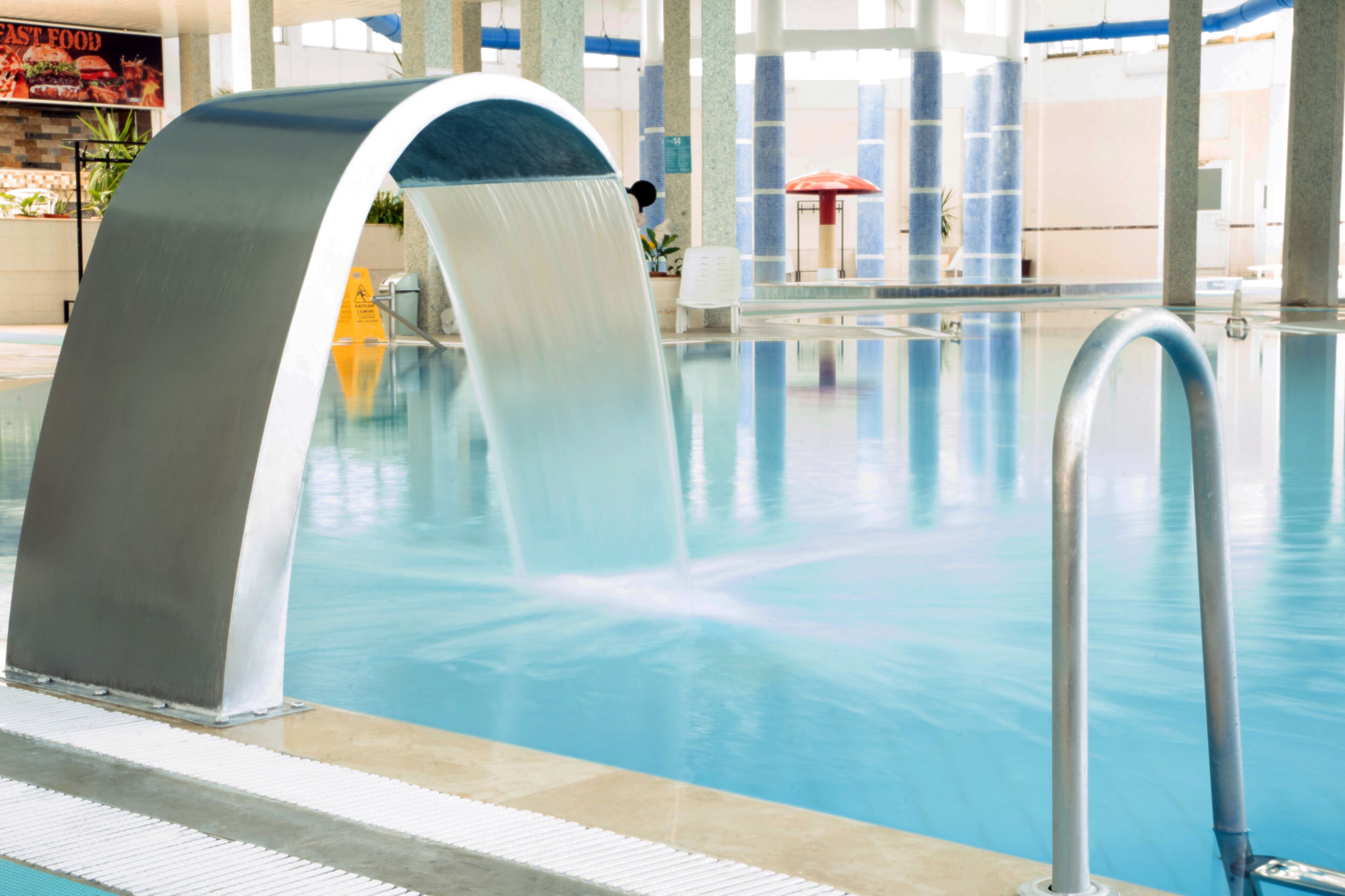 A swimming pool with a water feature, indoor seating area, and a fast food restaurant in the background.