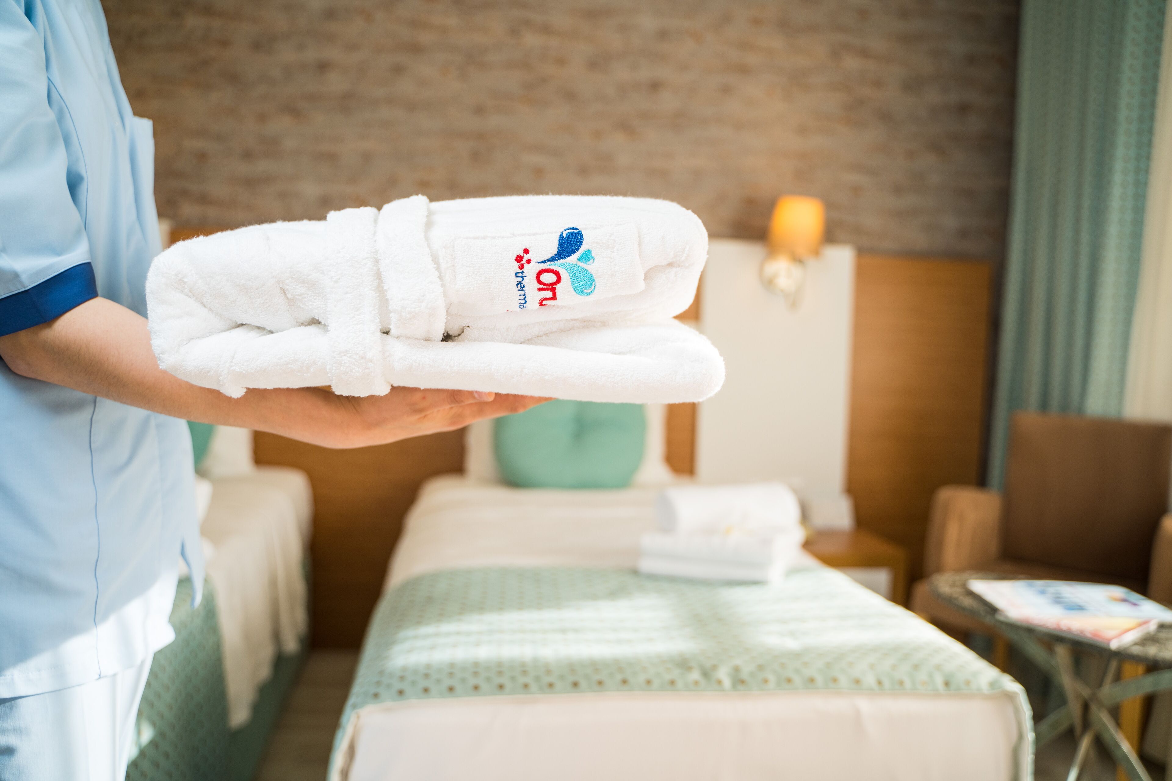 Une personne tenant une serviette blanche ornée d’un logo, dans une chambre d’hôtel équipée d’un lit, d’une chaise et d’une petite table.