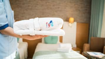 Une personne tenant une serviette blanche ornée d’un logo, dans une chambre d’hôtel équipée d’un lit, d’une chaise et d’une petite table.