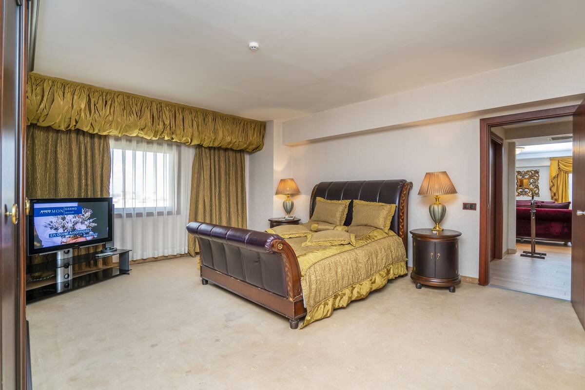 Une chambre spacieuse avec un grand lit, deux tables de chevet avec des lampes, un téléviseur posé sur un meuble et une vue sur une autre pièce grâce à une porte ouverte.