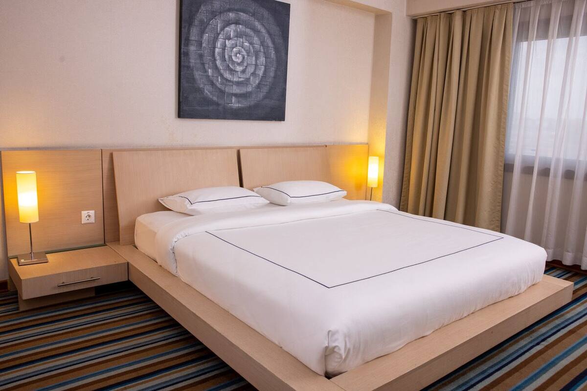 Une chambre d’hôtel avec un grand lit, deux tables de chevet avec des lampes, une œuvre d’art accrochée au mur et une fenêtre avec des rideaux.