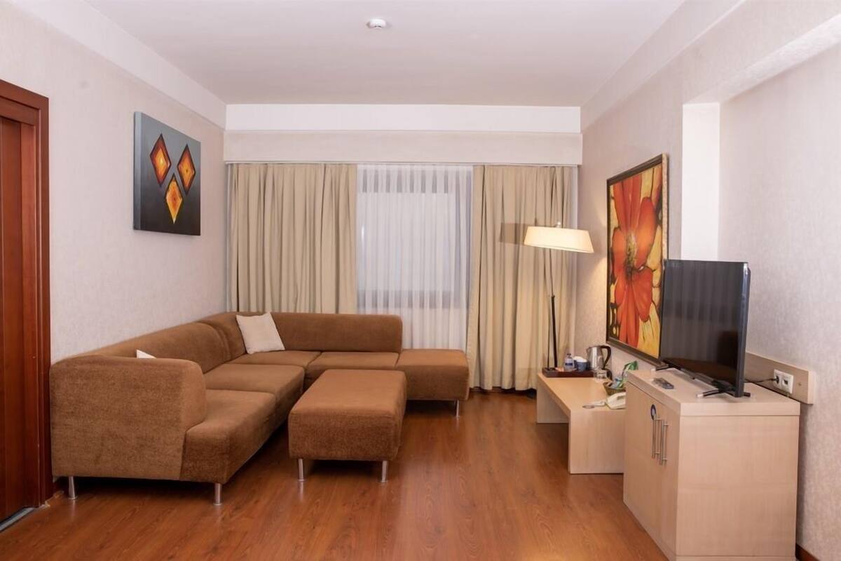 Une chambre d’hôtel moderne avec un canapé d’angle, un téléviseur posé sur un meuble, un bureau et un tableau accroché au mur.