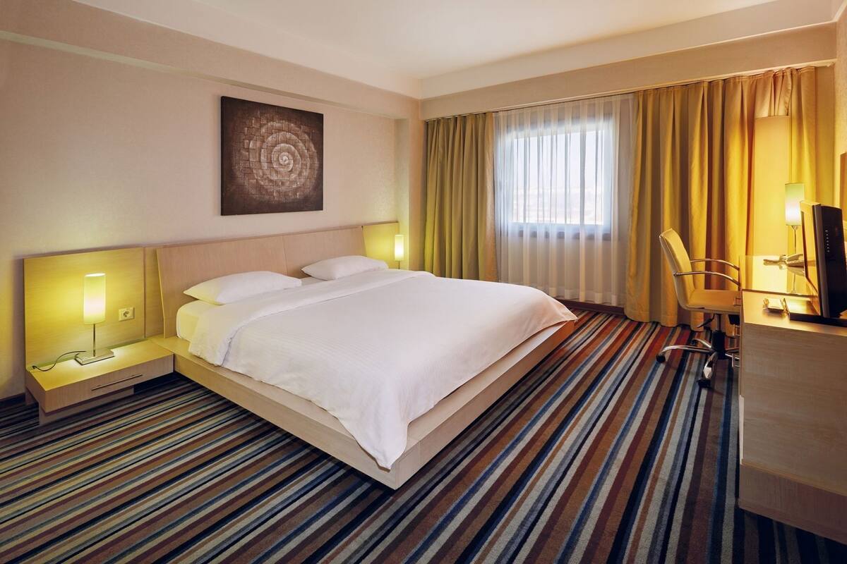 Une chambre d’hôtel avec un lit, des lampes de chevet, un bureau avec un ordinateur, une chaise et une grande fenêtre avec des rideaux.