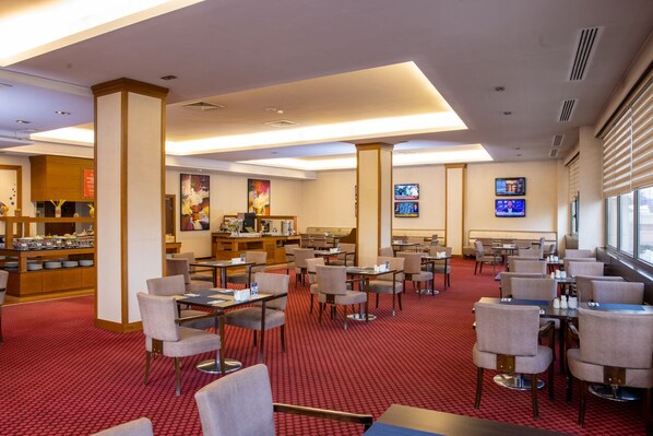 A hotel dining area with multiple tables and chairs, a buffet counter, and a television screen.