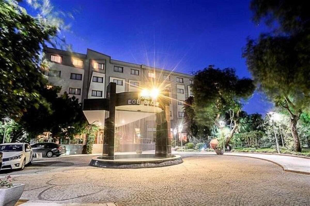 A hotel entrance with a circular fountain, a sign with the hotel's name, and a paved area with trees and parked cars.