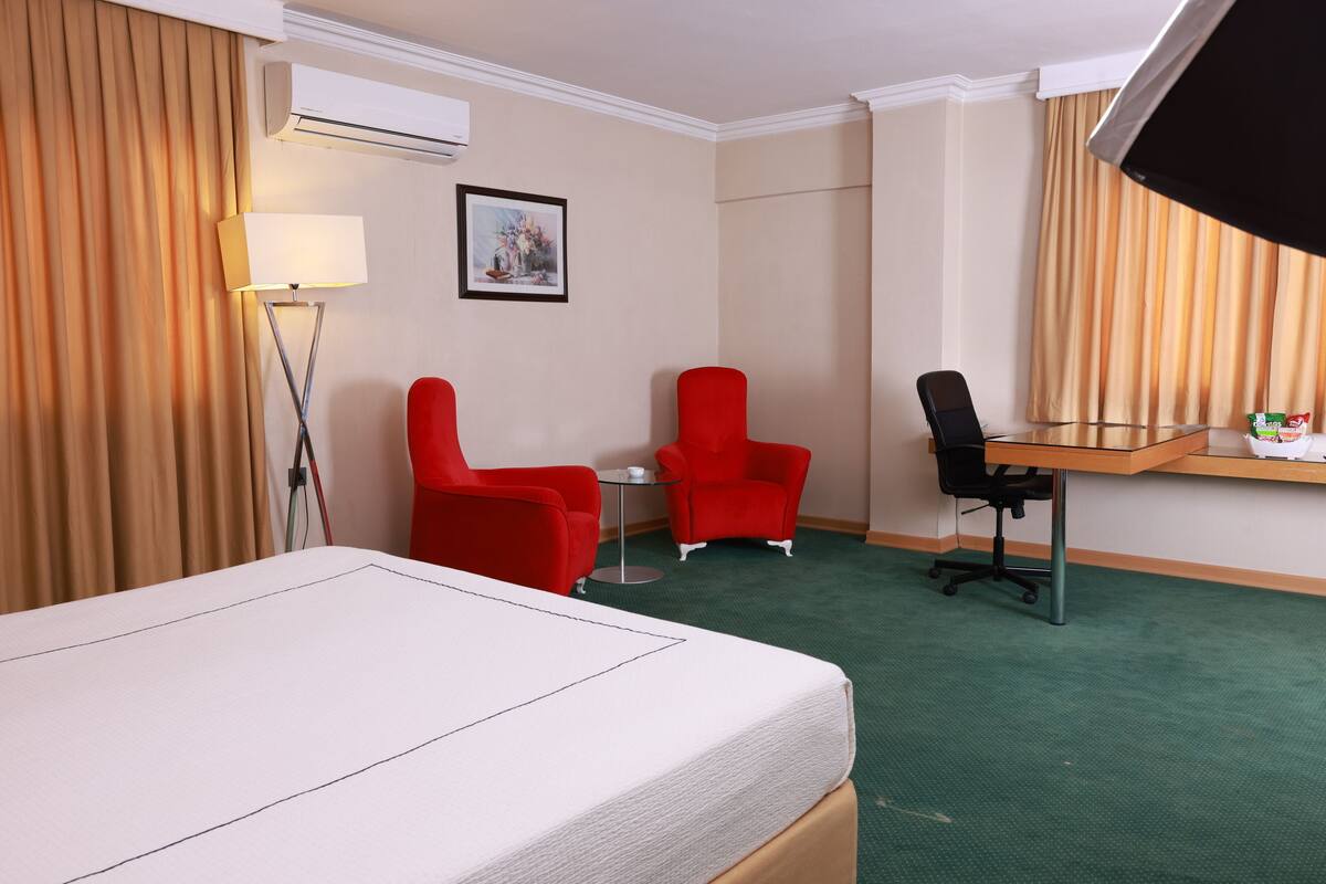 A hotel room with a bed, two red chairs, a small table, a desk, and a picture on the wall.