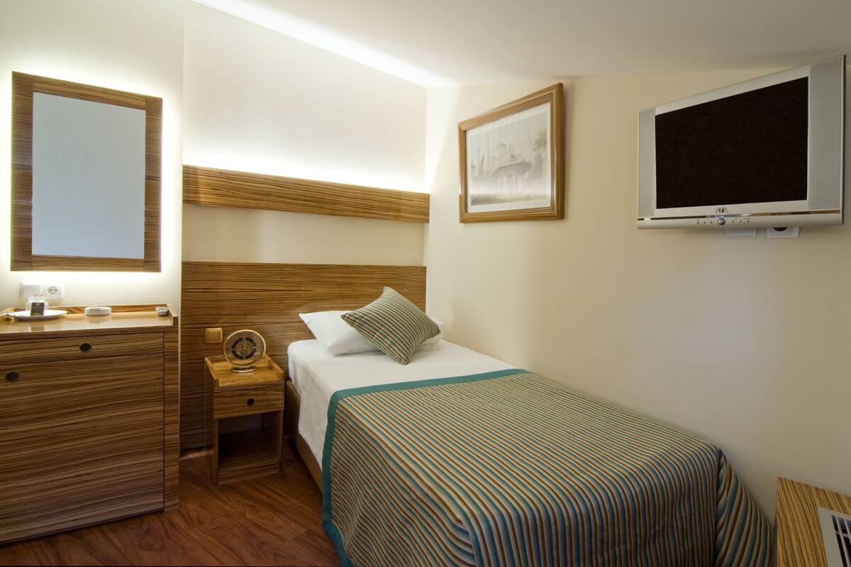 Ein Hotelzimmer mit einem Holzschrank, einem Nachttisch mit Lampe, einem Bett mit gestreifter Decke, einem gerahmten Bild an der Wand und einem an der Wand befestigten Fernseher.