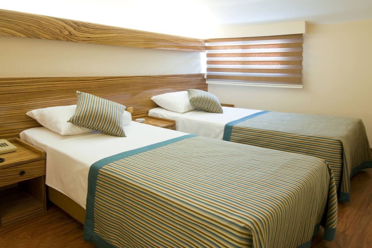 Ein Hotelzimmer mit zwei Betten, Holz-Kopfteilen, gestreiften Bettdecken und einem Fenster mit Jalousien.
