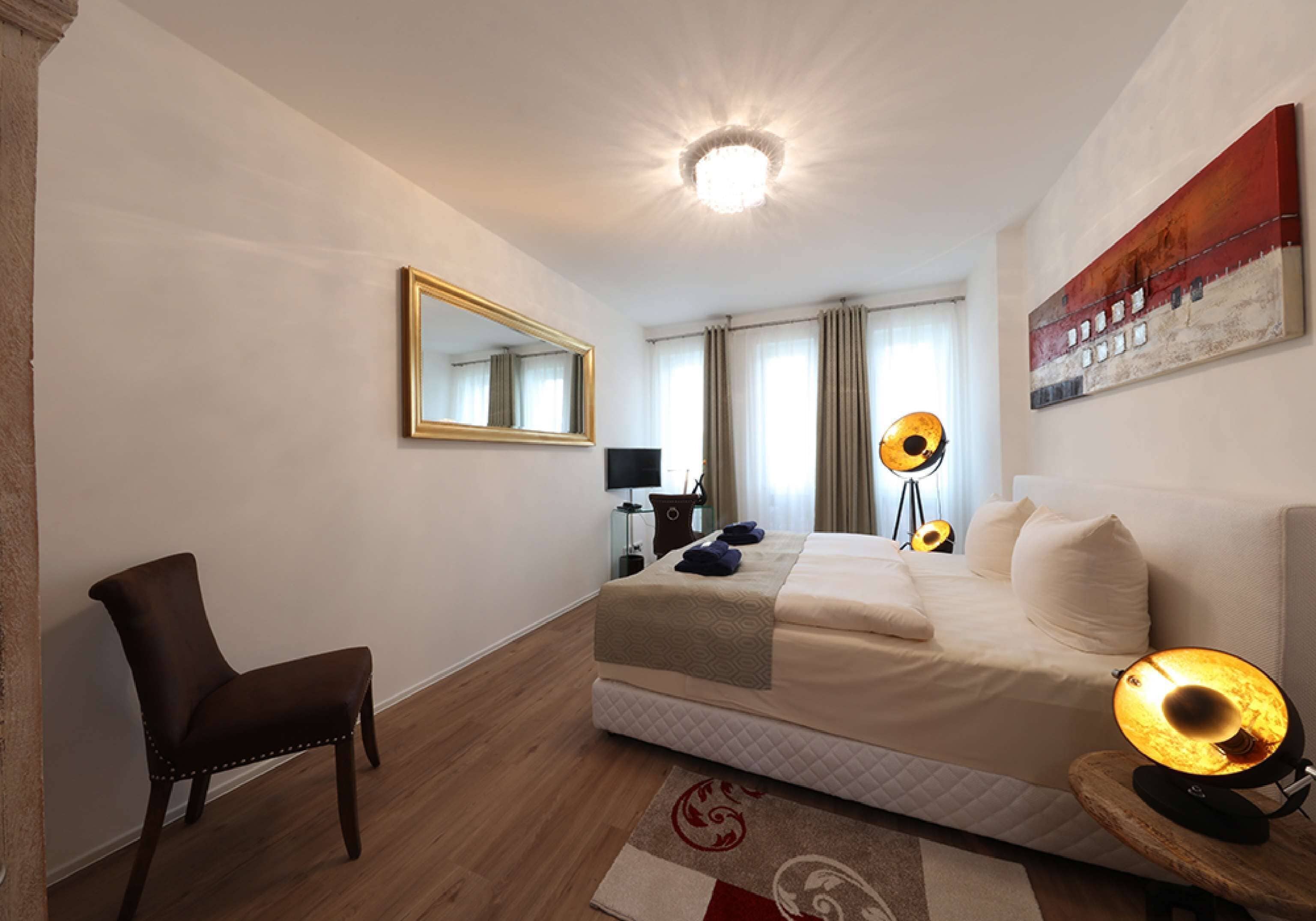 A hotel room with a bed, a chair, a desk with a lamp, a mirror, and a painting on the wall.