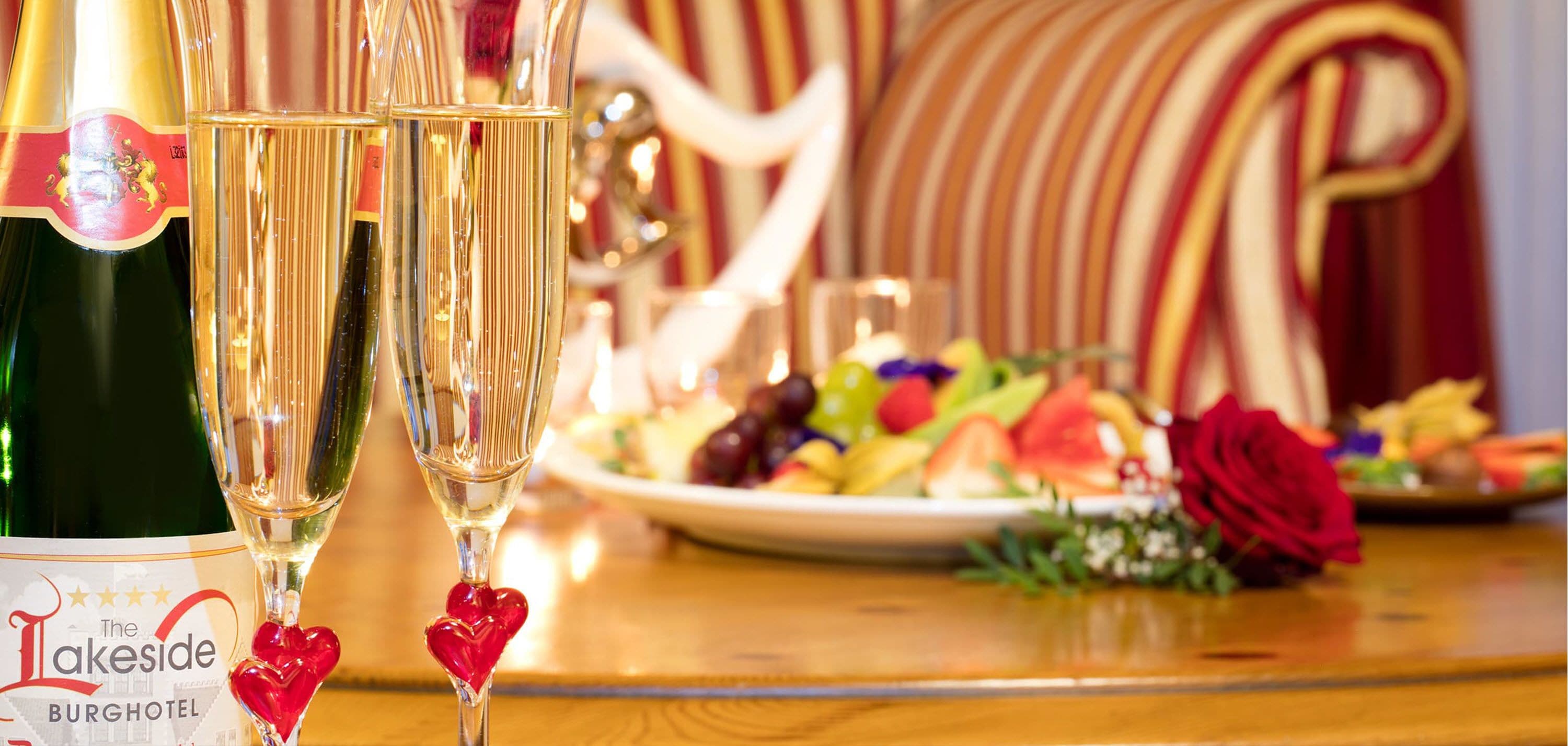 A bottle of wine and two glasses of champagne with heart-shaped decorations, a plate of assorted fruits, and a red rose on a wooden table.
