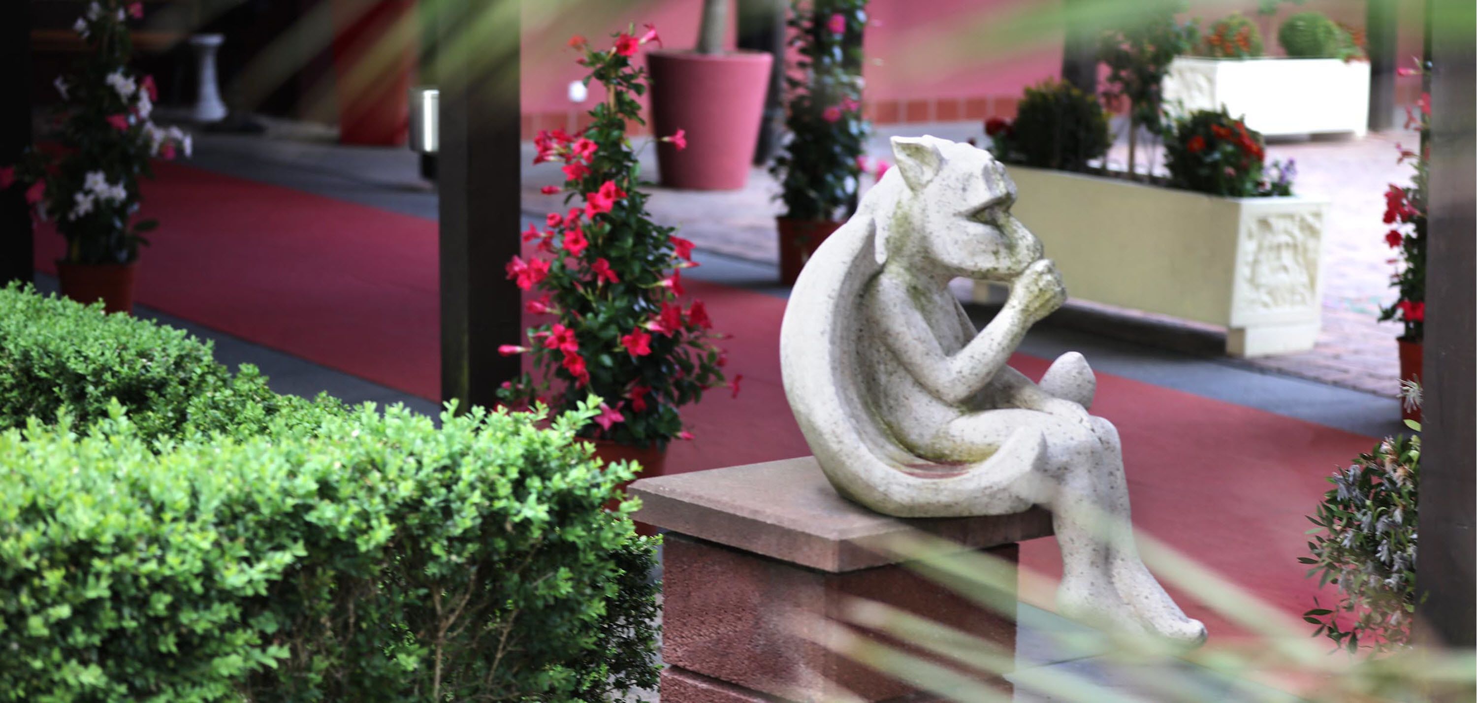 Een stenen sculptuur van een zittend figuur in een tuin, met potplanten en een rode loper.