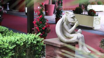 Een stenen sculptuur van een zittend figuur in een tuin, met potplanten en een rode loper.