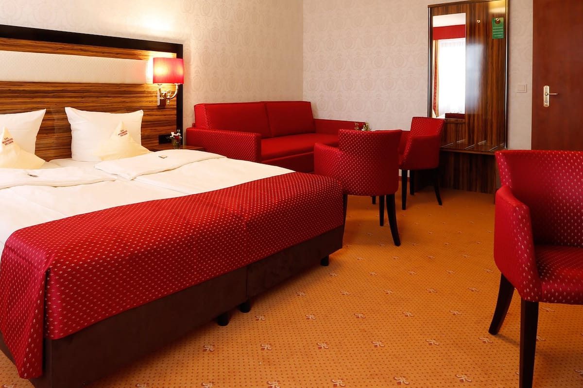 Ein Hotelzimmer mit einem großen Bett, roten Polstersesseln, einem Holzkopfende und einer rot gemusterten Bettdecke.