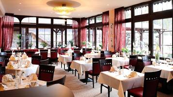 A restaurant with multiple tables, red chairs, and large windows with curtains.