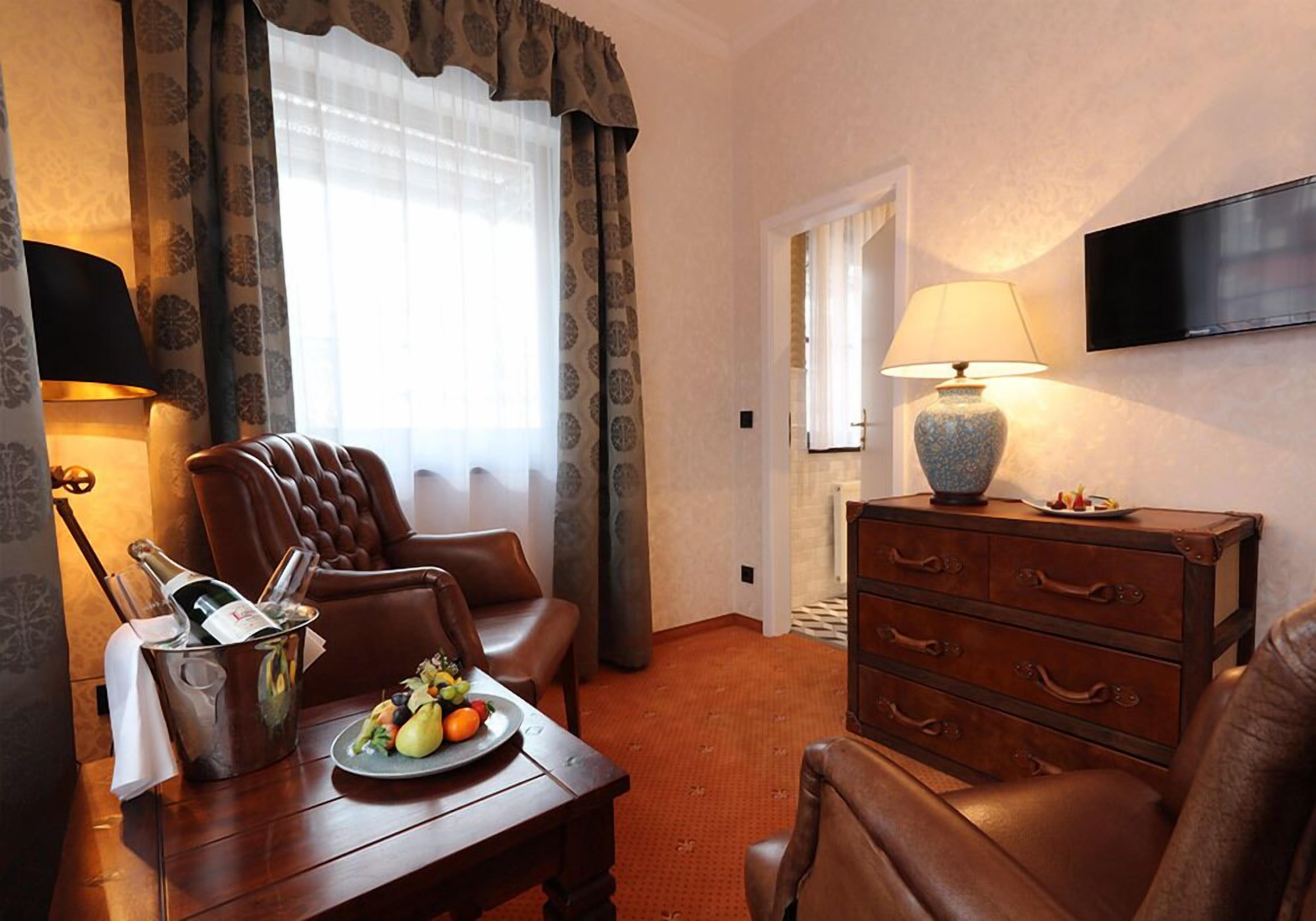 A hotel room with a leather armchair, a wooden table with a fruit plate, a bottle of champagne, and a lamp.